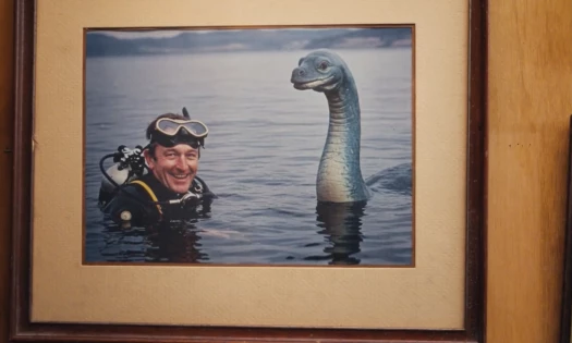 A man is shown happily posing with the Loch Ness monster