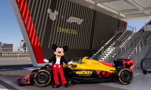 Disney's Mickey Mouse poses with a "Mickey and Friends" racecar with the two brands' logos and "COMING 2026" in the background