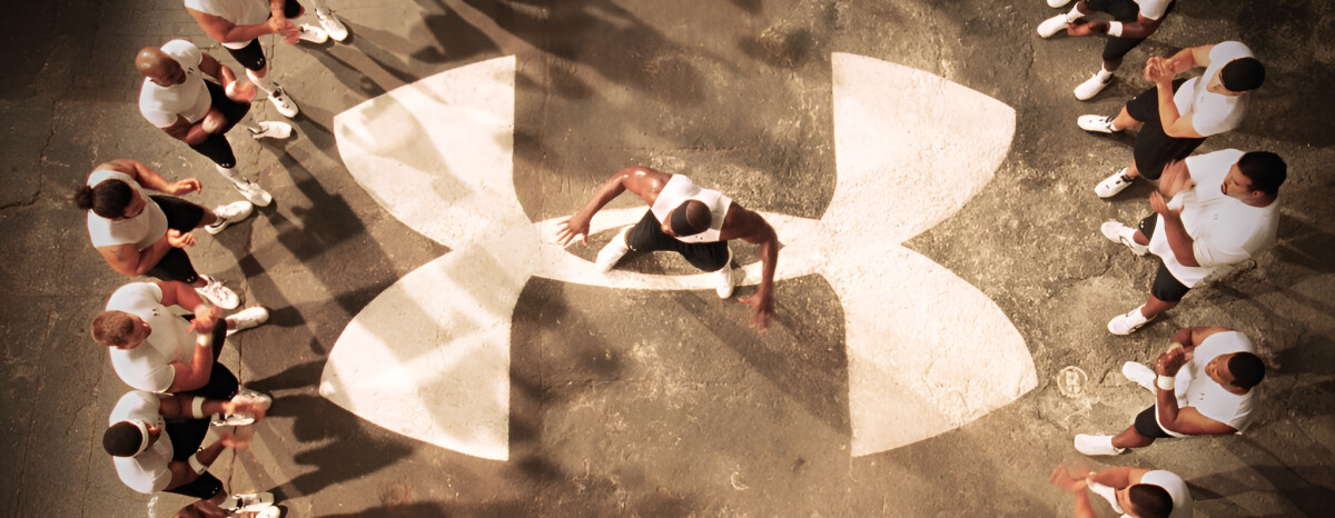 Eric "Big E" Ogbogu standing at the center of a big Under Armour logo with athletes clapping surrounding him from the brand's first national TV ad, "Protect this House"
