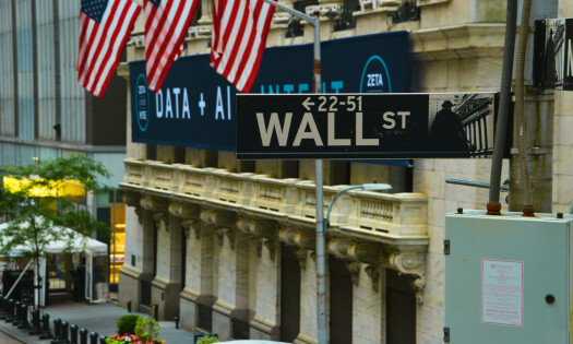 A Wall Street sign with the stock exchange building in the background with American flags