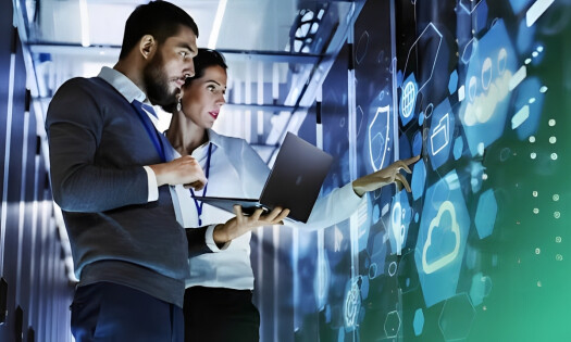 Two professionals, a bearded man and a woman in business attire, collaborate in a dimly lit server room. The man points at floating holographic icons representing cloud computing, security locks, and data symbols, while the woman holds an open laptop. The