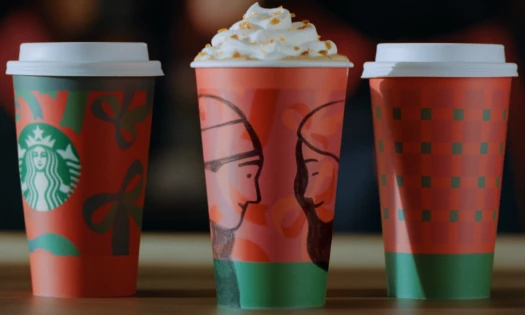 Three Starbucks cups sitting on a table, showing this year's holiday-themed illustrations