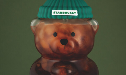A photo of the Starbucks "Bearista Cold Cup" filled with coffee, which looks like a transparent teddy bear holding a coffee cup and wearing a green beanie