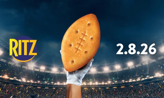 A gloved hand is holding a football-shaped RITZ cracker, in front of a backdrop showing a football stadium