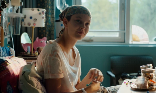 A young female cancer patient sitting inside a bedroom, looking towards the camera and smiling