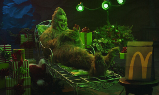 The Grinch sitting on a lawn chair outside at nighttime, holding a phone and laughing, surrounded by Christmas presents, a tree and a McDonald's bag