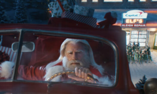 John Travolta dressed as Santa sitting inside a red car, in front of a Christmas-themed scene