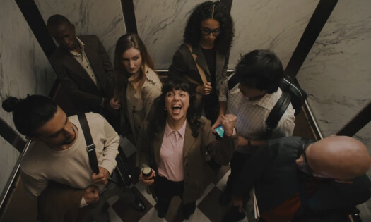 A group of people are shown excitedly celebrating football inside an elevator, with a woman standing in the center holding a bottle of Coke