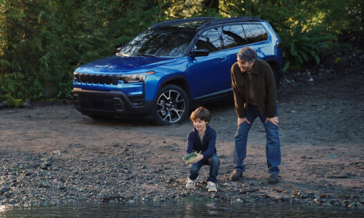A father and son releasing Big Mouth Billy Bass in the river with a blue 2026 Jeep Cherokee Hybrid in the background