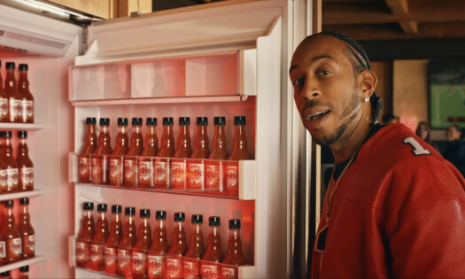 Rapper Ludacris standing beside a fridge filled with bottles of Frank's RedHot sauce