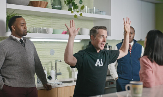 A man wearing a shirt with the text "AI" on the front is standing inside a workplace kitchen with his arms in the air, surrounded by confused-looking colleagues
