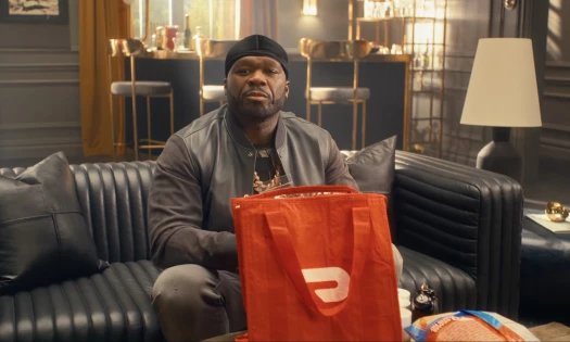 Rapper 50 Cent sitting on a couch and smiling, with a DoorDash bag sitting on a table in front of him