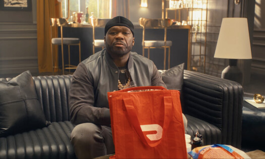 Rapper 50 Cent sitting on a couch and smiling, with a DoorDash bag sitting on a table in front of him