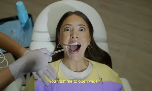 A woman lying on a dentist's chair with her mouth open, with the text "You want me to open wide?" shown at the bottom of the screen