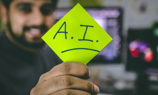 A man holding a note that says "A.I." while computer screens can be seen in the background