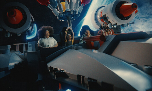 A promotional photo for the LEGOLAND Galacticoaster, showing four people, including the spot's child star, riding with their hands in the air
