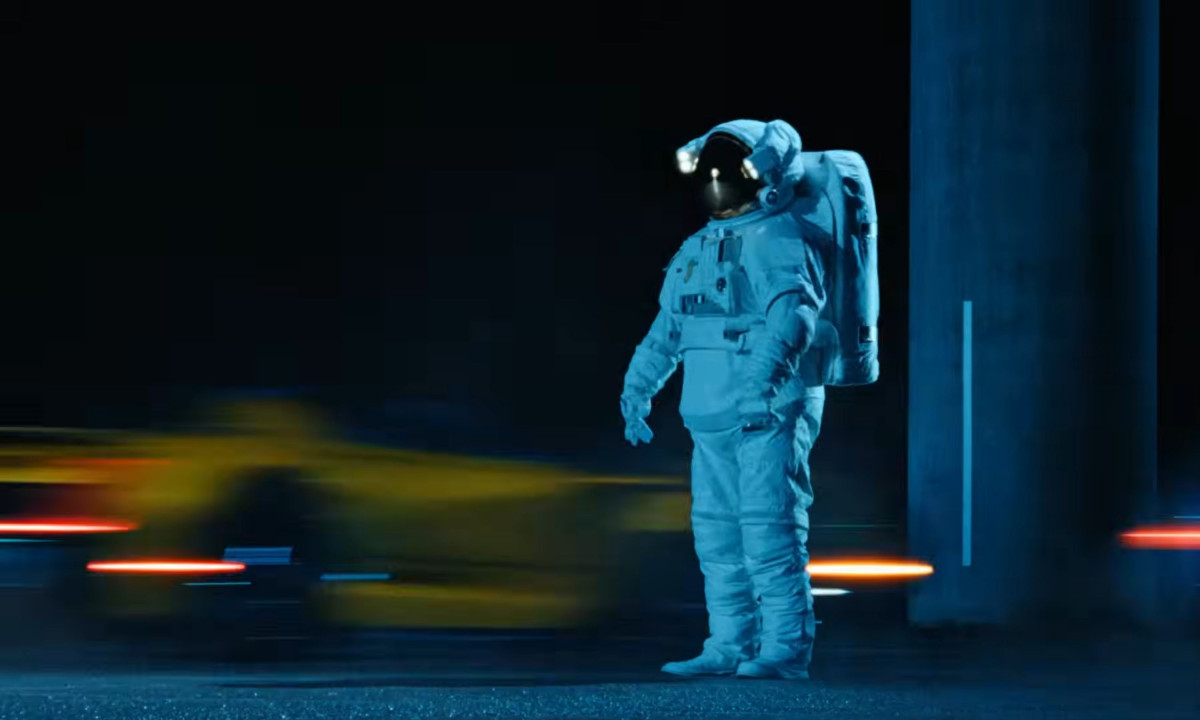 An astronaut in a full white suit standing on a road as a yellow car speeds by in a blur