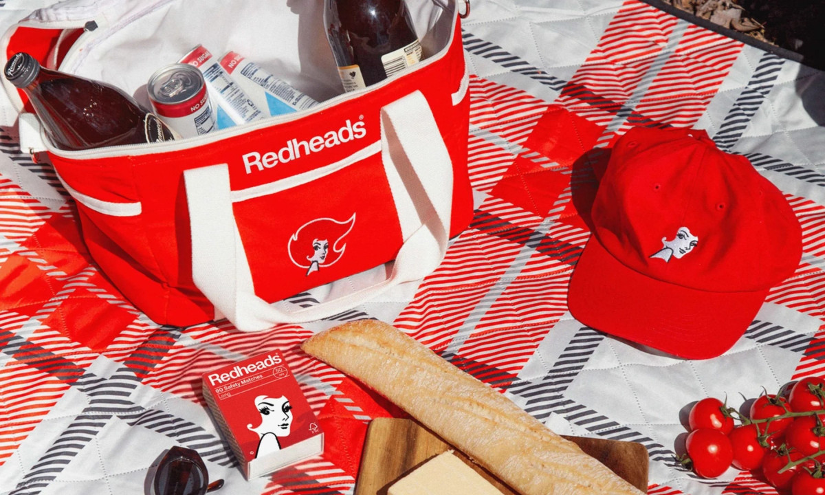 A red cooler bag and matching baseball cap featuring the Redheads logo placed on a red and white checkered picnic blanket with bread and tomatoes.