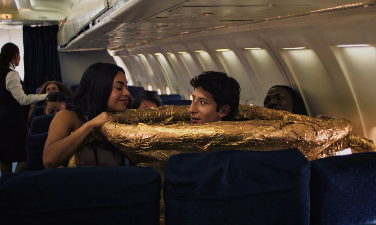 A man wearing a massive golden trophy costume sits in a crowded airplane cabin while the passenger next to him smiles.