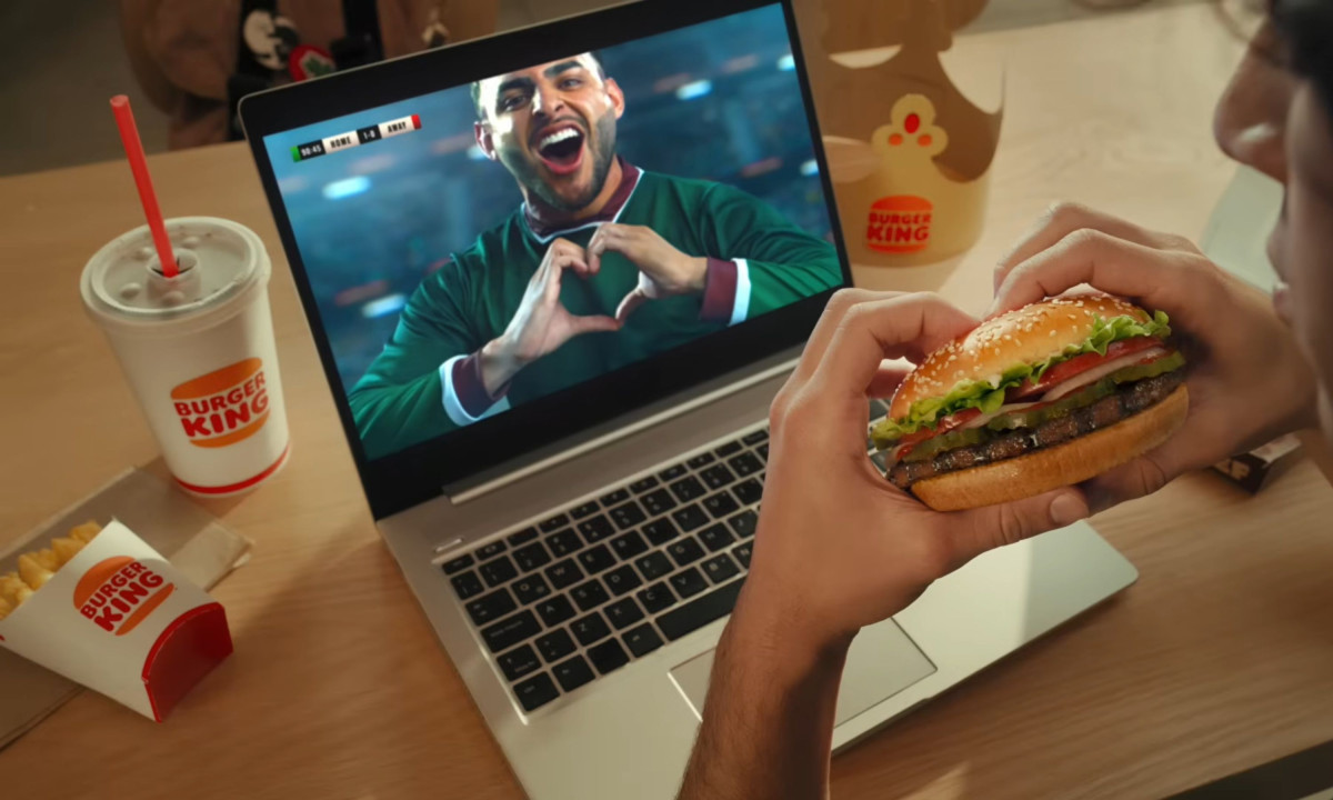 A person holds a Whopper while watching a soccer player form a heart on a laptop.