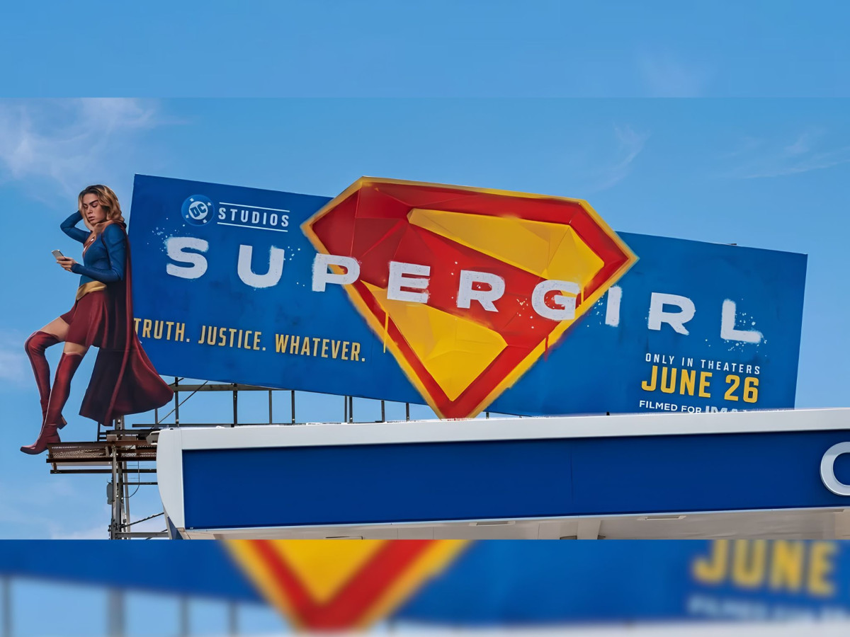Supergirl leaning on a billboard scrolling on her phone.