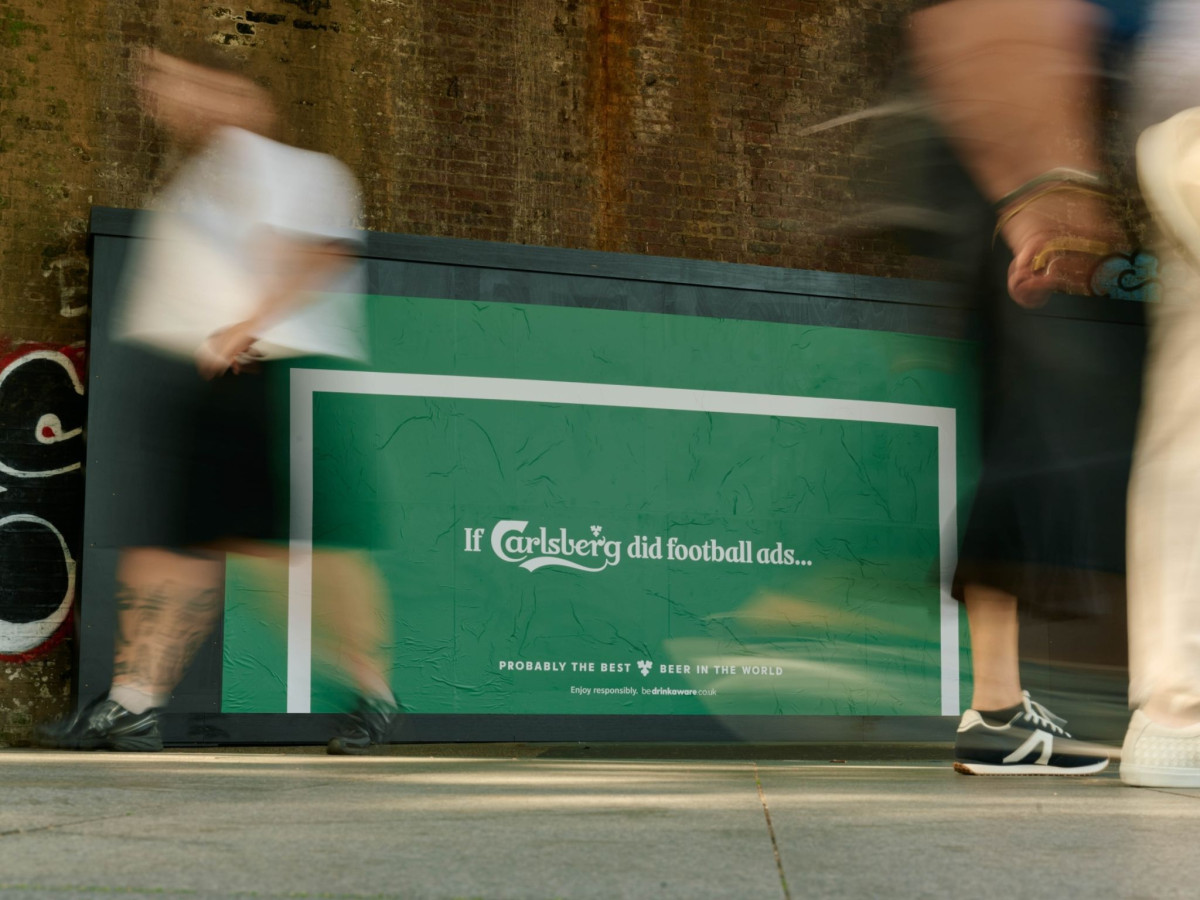 Blurred pedestrians walking past a green Carlsberg goal poster.