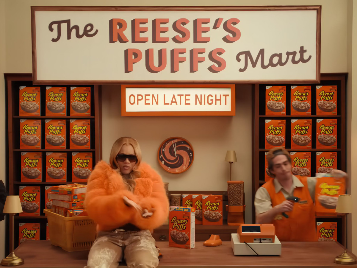 Rapper GloRilla in an orange fur coat sitting on a checkout counter inside The Reese's Puffs Mart.