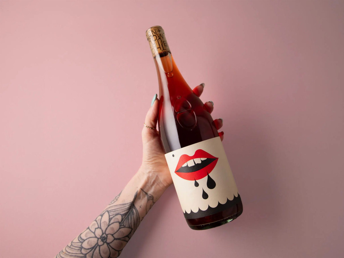 Tattooed arm holding a bottle of wine featuring a graphic red lips label.