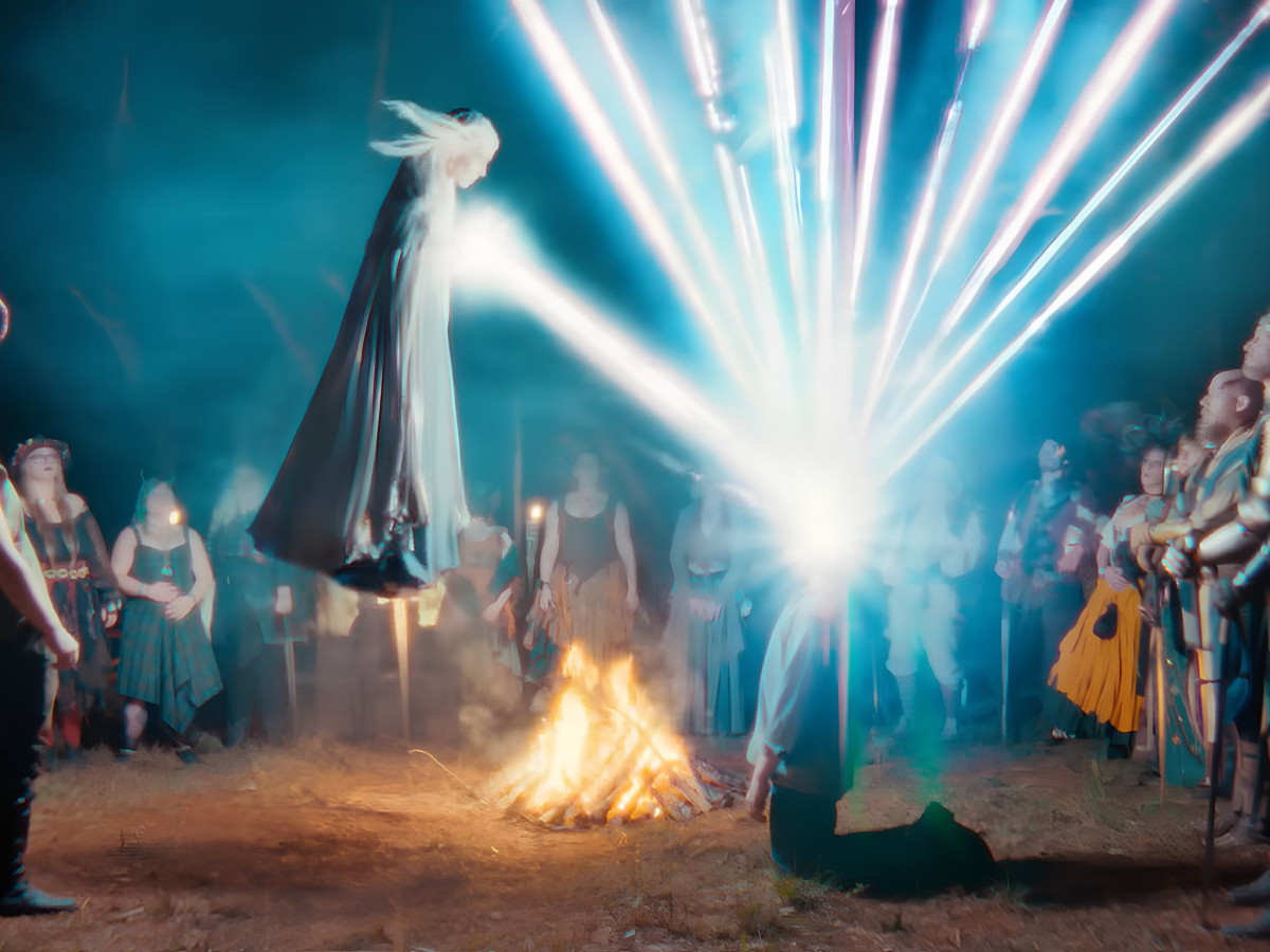 Floating cloaked figure emitting a beam of light onto a kneeling person by a campfire.