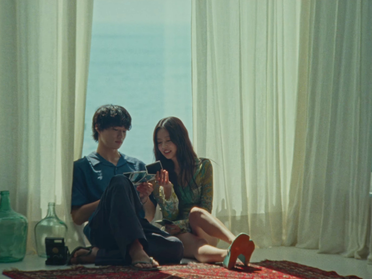 Jennie and Nah Kai seated on a red Persian rug on a white floor, looking at Polaroid photos together, sheer white curtains framing an ocean view behind them, large green glass bottle to the left, vintage camera on the floor