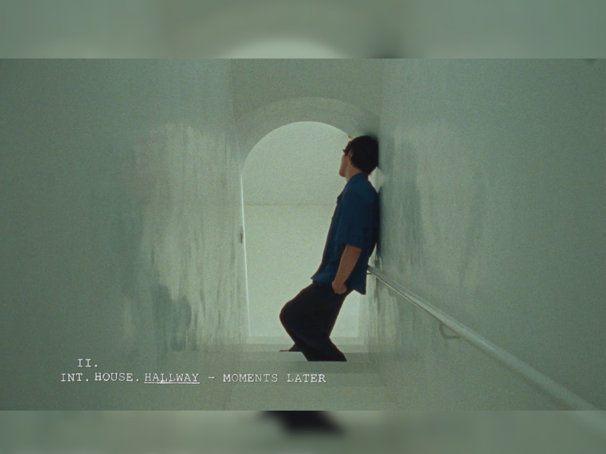 Nah Kai in silhouette leaning against a pale green hallway wall, arched doorway behind him with bright white light flooding through, scene slug reading "II. INT. HOUSE. HALLWAY — MOMENTS LATER" in typewriter font at bottom left, heavy film grain throughou