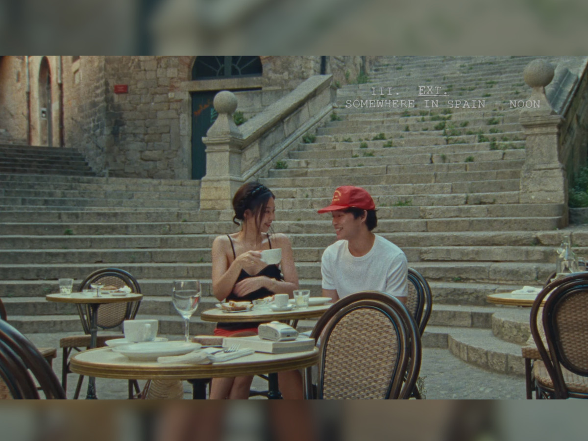 Jennie and Nah Kai laughing at a bistro table in an outdoor Spanish café, ancient stone staircase rising behind them, Jennie in black camisole with red skirt, Nah Kai in white tee and red cap, scene slug "III. EXT. SOMEWHERE IN SPAIN — NOON" upper right