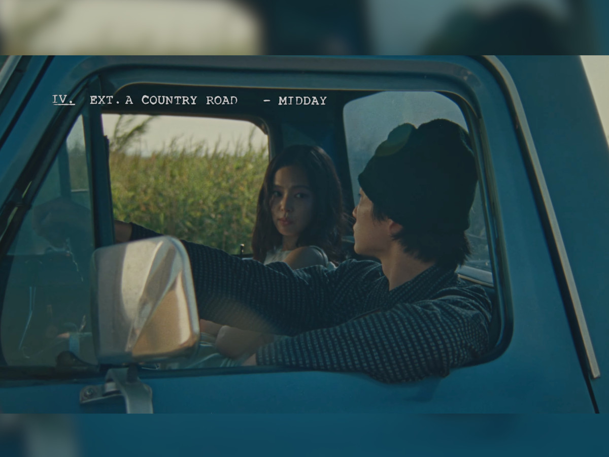 Jennie and Nah Kai inside a vintage blue truck cab, scene slug "IV. EXT. A COUNTRY ROAD — MIDDAY" upper left, Jennie looking directly at camera through the window frame, desaturated cool blue-green grade, cornfield visible through rear window