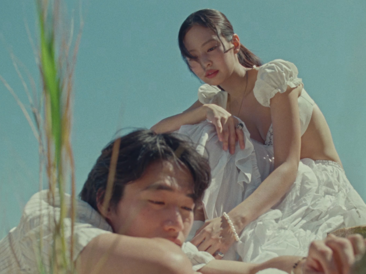Low-angle shot looking up at Jennie in a white ruffled puff-sleeve crop top seated above Nah Kai who lies in her lap, wild grass breaking the left foreground, clear blue sky as backdrop, warm directional sunlight casting shadows across both figures