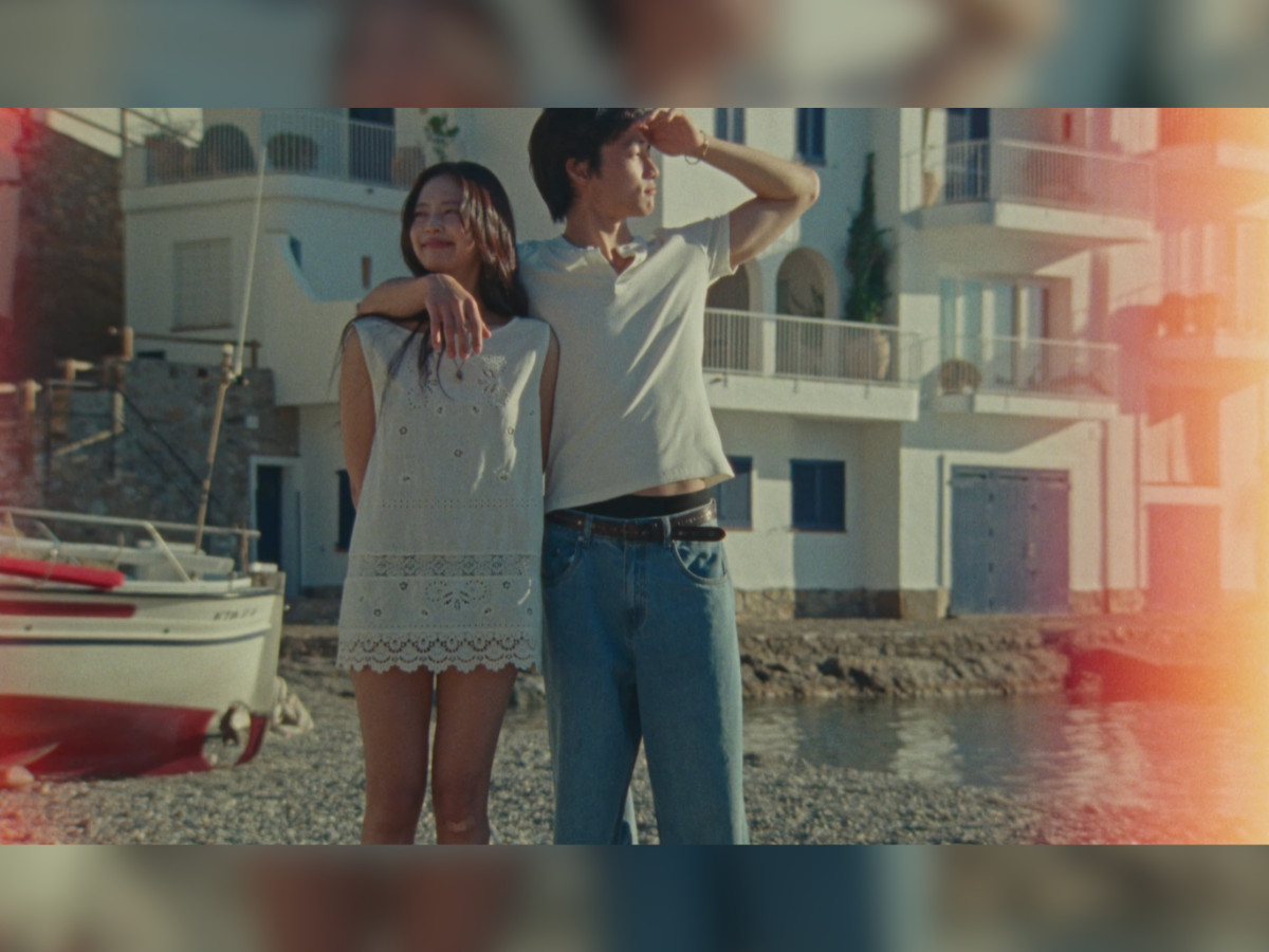 Jennie in white broderie anglaise mini dress standing beside Nah Kai at a harbour's edge, his arm around her shoulder, Mediterranean white and blue buildings behind them, red and white boats moored to the left, strong orange lens flare bleeding into the r