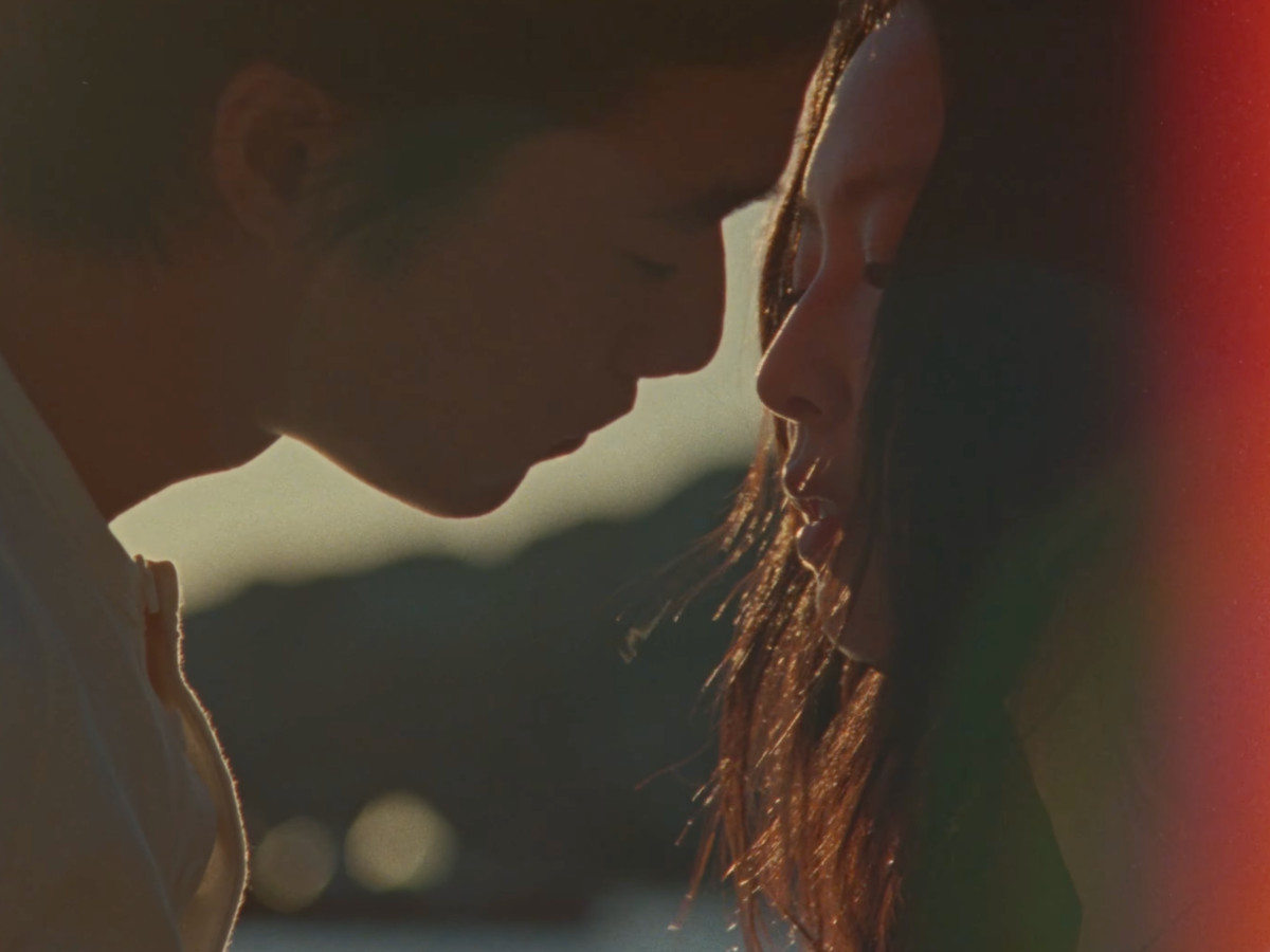 Extreme close-up of Jennie and Nah Kai facing each other, foreheads nearly touching, backlit by golden hour sun that halos their hair, large circular bokeh lights visible in background, deep red-orange lens flare bleeding into right side of frame, both fa
