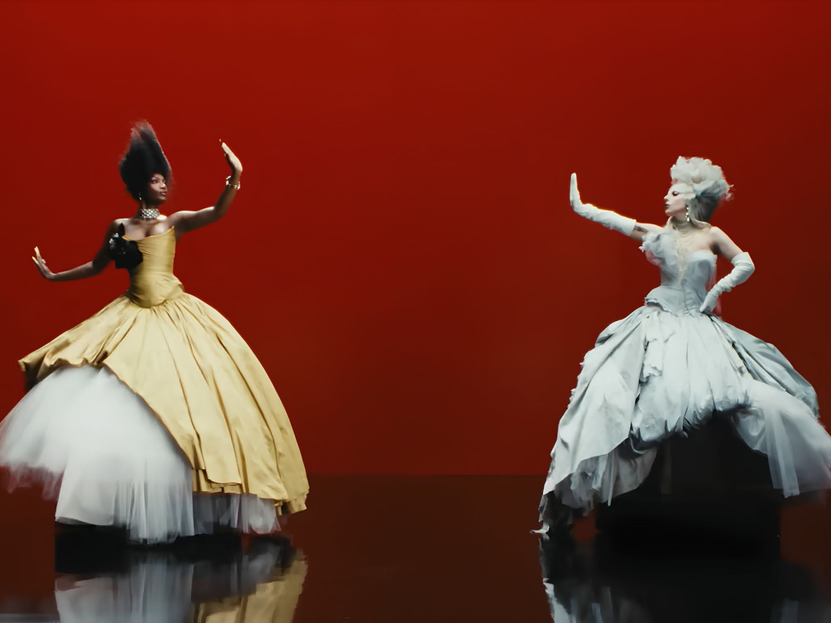 Lady Gaga and Doechii posing in voluminous, avant-garde gowns against a stark red background.