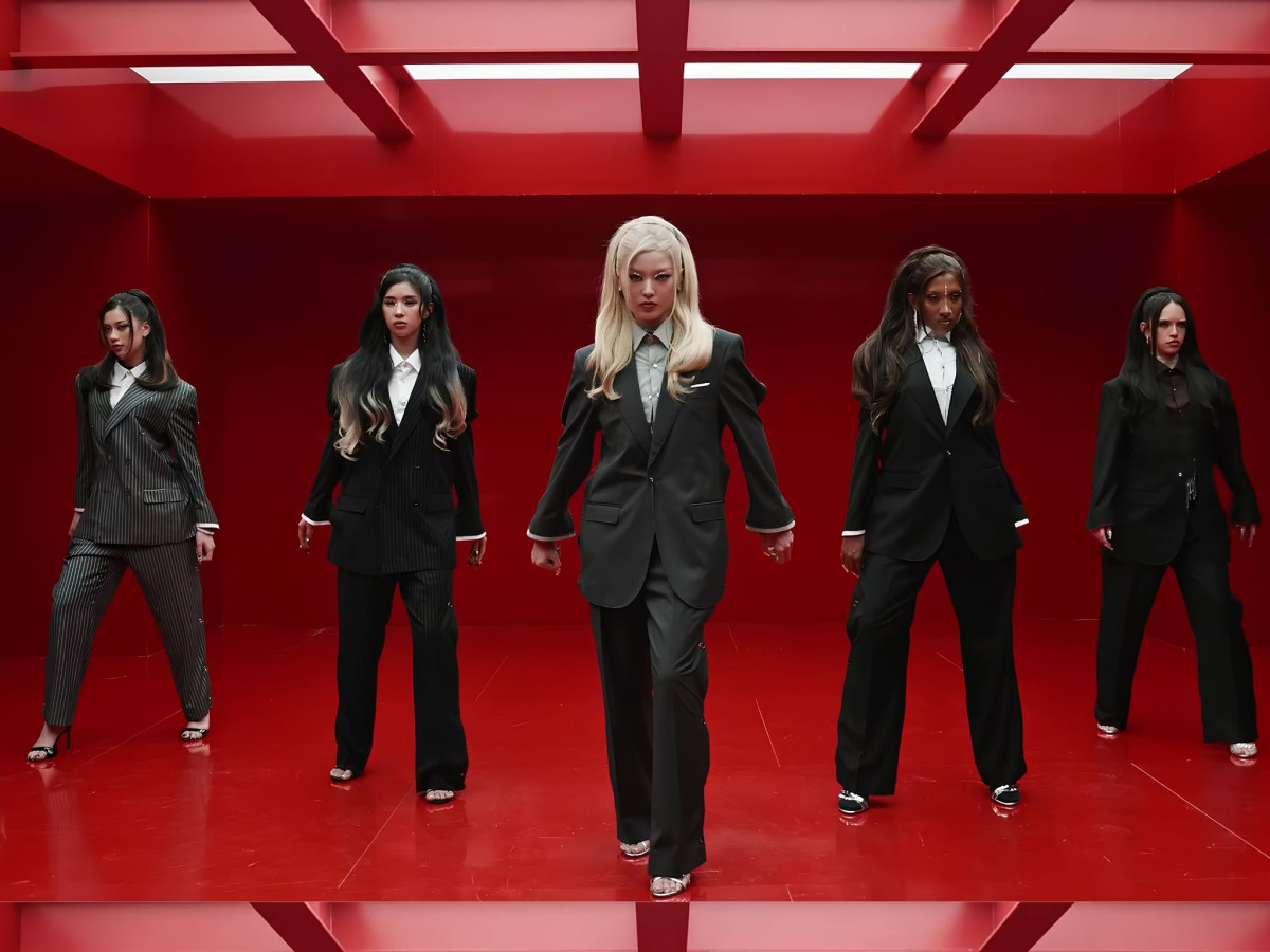 Five KATSEYE members wearing tailored, oversized suits standing in a stark, bright red geometric room.