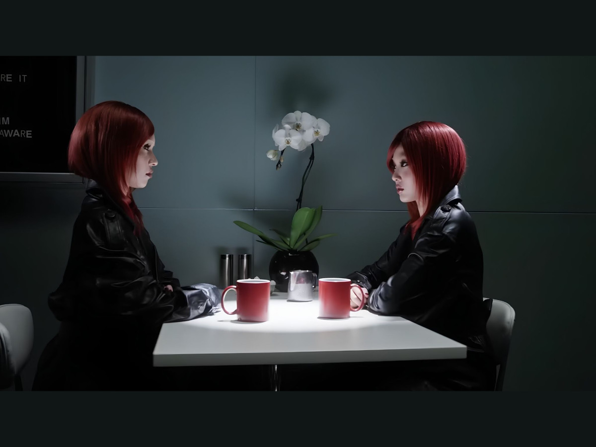 Two women with red bob haircuts wearing black leather jackets sitting across from each other at a white table in a dimly lit room.