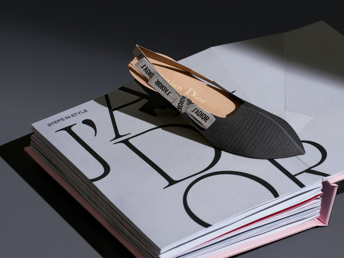An opened pop-up book displaying a 3D rendition of a single Dior black pump