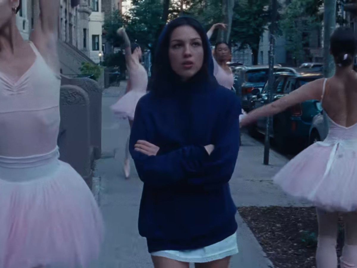 Olivia Rodrigo in a blue hoodie walking past ballerinas.