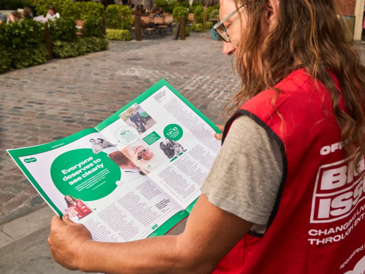 Vendor in a red vest reading an oversized magazine spread.