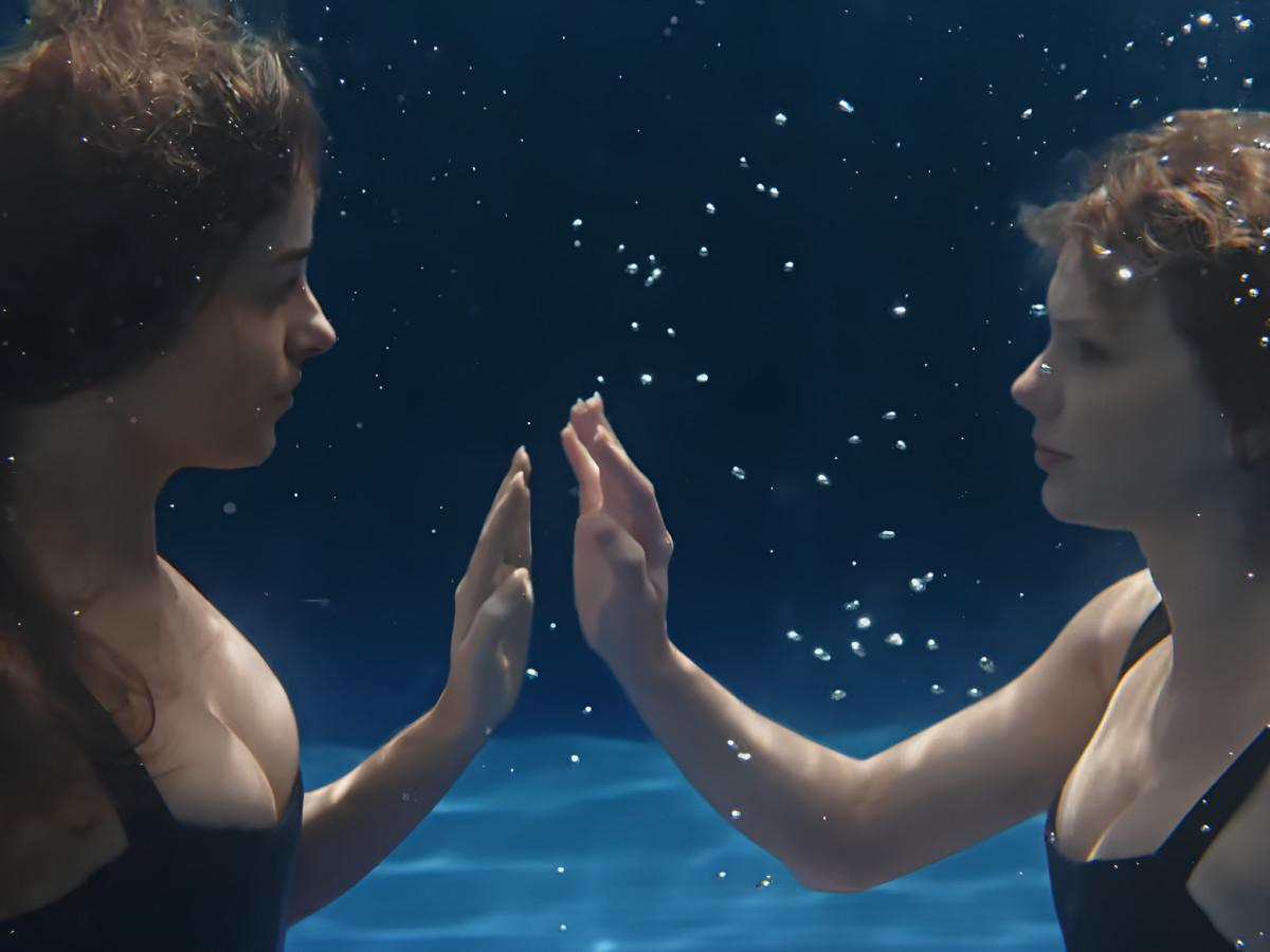 Two women underwater touching hands through glass.