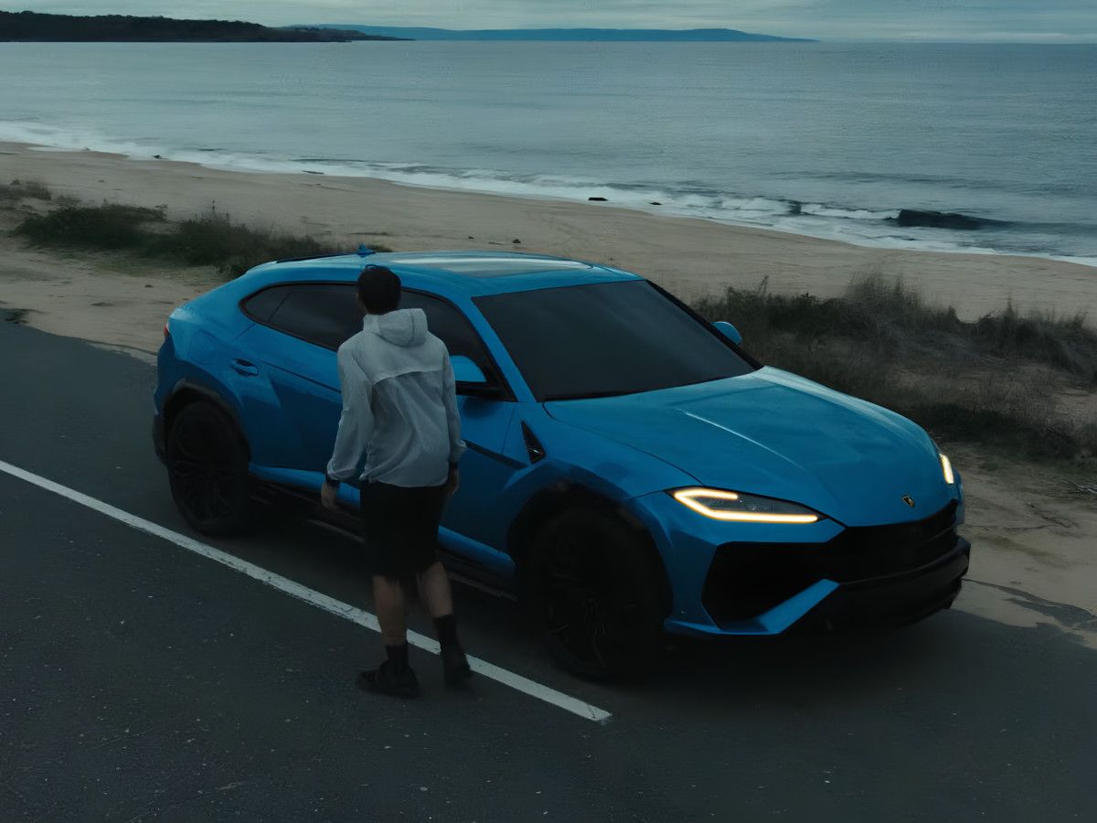 Man walking towards a blue Lamborghini parked on a coastal road beside a beach.
