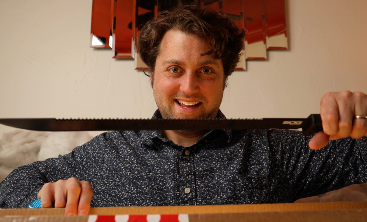 A man smiling while holding a serrated knife in front of a cardboard box.