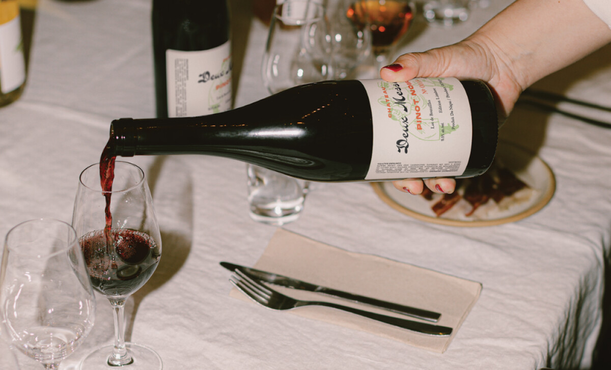 A person's hand pours red wine from a bottle of Deux Messieurs Pinot Noir into a glass by Haus Haus Design.