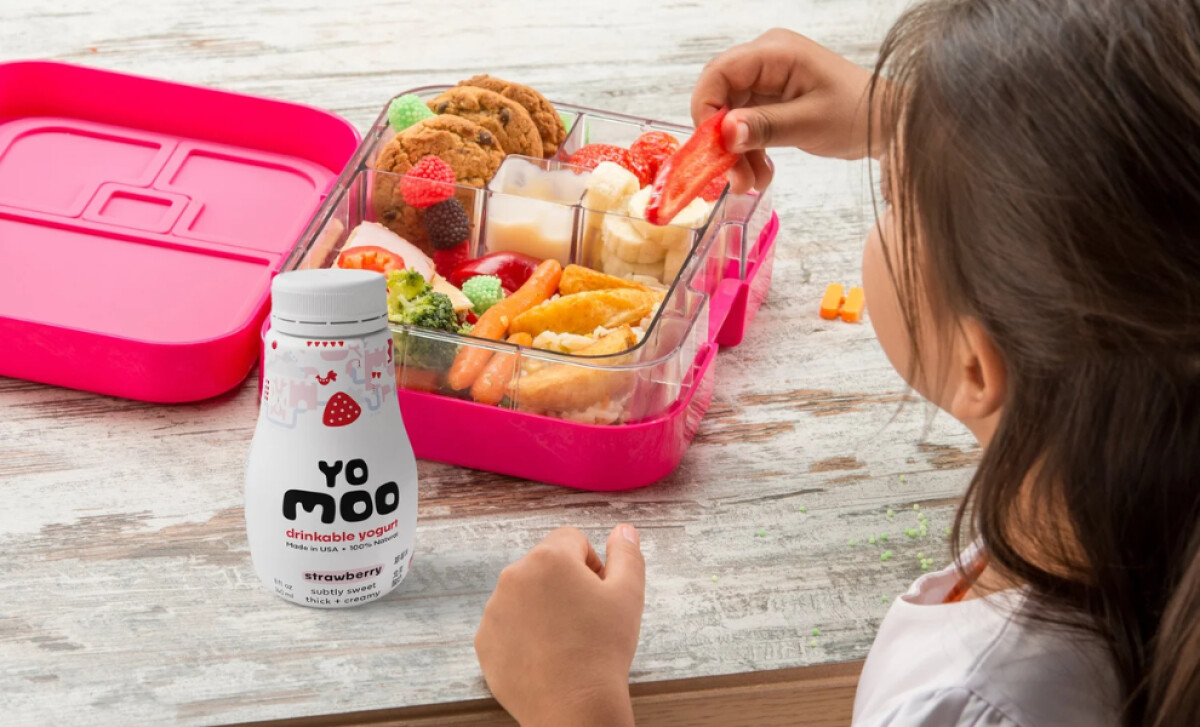 A young girl eats from a pink bento box, with a bottle of Yomoo yogurt by BLC Design.