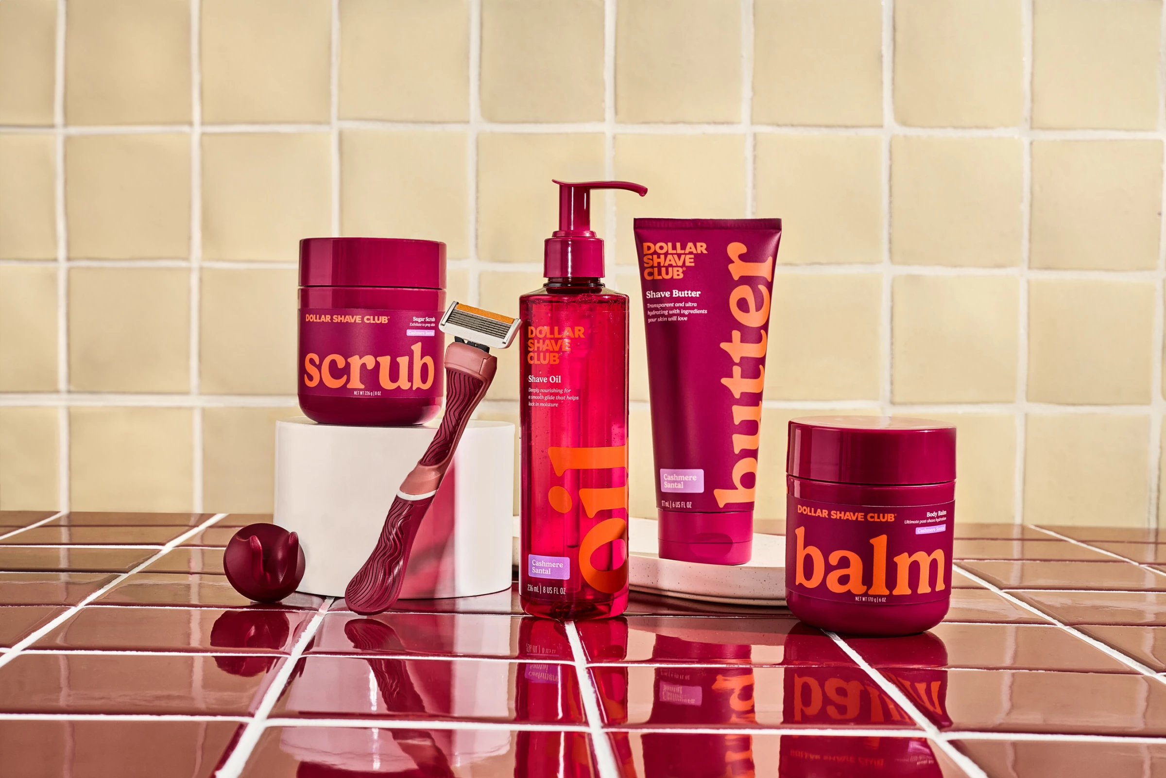 Dollar Shave Club New Women's Line Dollar Shave Club women’s shaving products including razor, shave oil, butter, scrub, and balm displayed on tiled bathroom surface in bold maroon packaging.