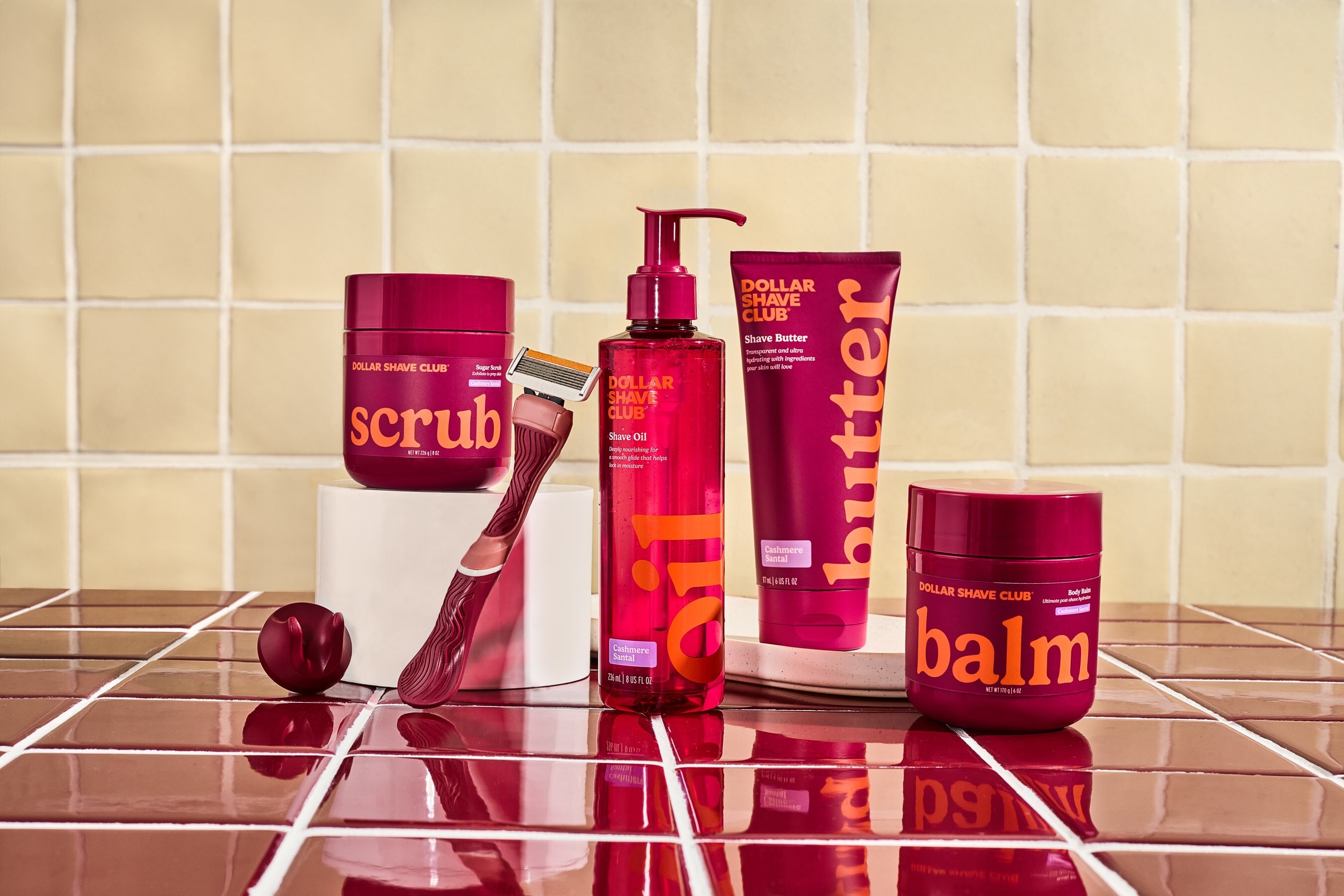 Dollar Shave Club women’s shaving products including razor, shave oil, butter, scrub, and balm displayed on tiled bathroom surface in bold maroon packaging.