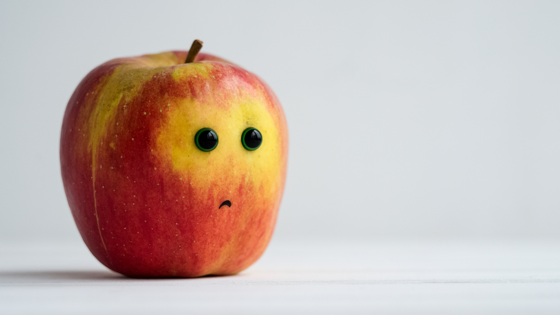 An image of a red and yellow apple with oversized green eyes and a small downturned mouth, sitting on a white surface against a plain background.